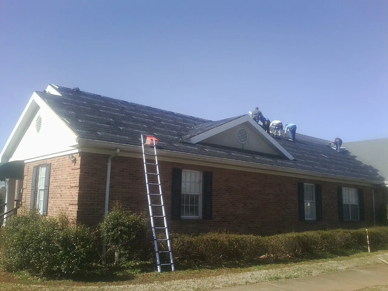 Crew tearing shingles off a brick ranch roof with a ladder set — Metal Roof Repair work by our crew in Footville