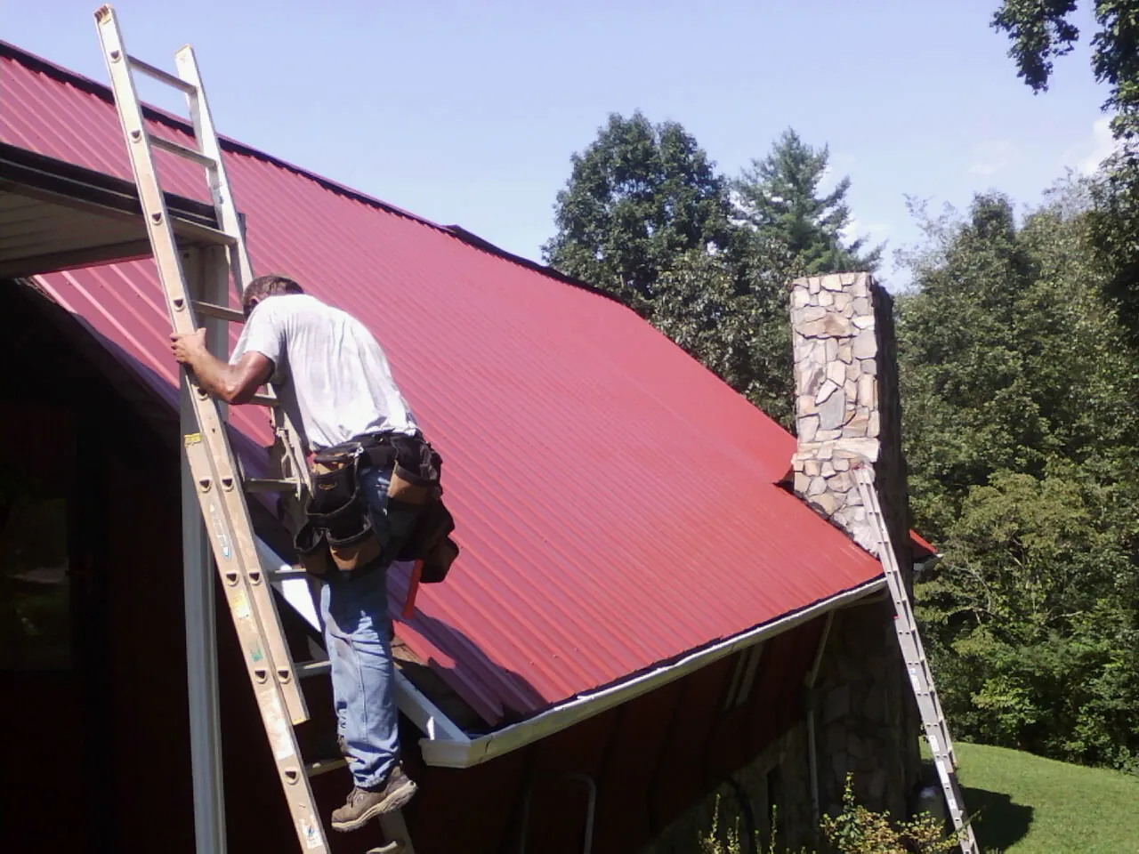 Roofer on a ladder beside a red metal roof and stone chimney — Soffit & Fascia Repair by Roofing Brothers in Footville